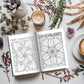 Coloring book open to pages with floral designs and humorous text, surrounded by candles, plants, and a cup of coffee on a white surface.