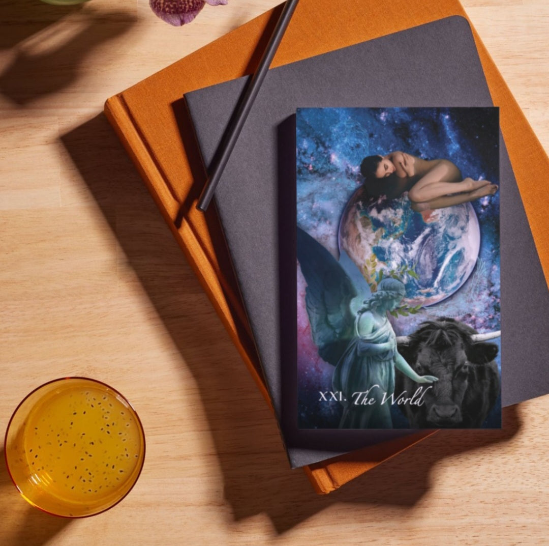 Tarot card titled 'XXI. The World' on a wooden surface with books and a cup.