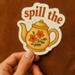 Sticker with a teapot design and 'spill the' text held by a hand against a brown background
